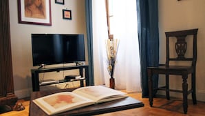 A living room with a flat-screen TV on a stand, a wooden chair, a table with an open book, and a vase with dried flowers.