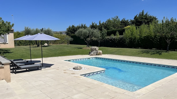 A rectangular swimming pool with a tiled edge, surrounded by a paved patio. There are lounge chairs and a blue umbrella on the patio. - Villa in Provence (Cavaillon)
