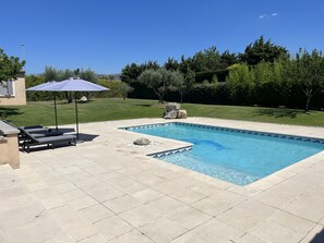 Una piscina rectangular amb un vorejador de rajoles, envoltada d'un pati pavimentat. Hi ha gandules i un parasol blau al pati.