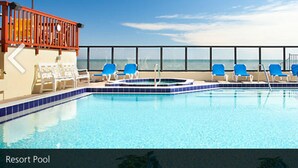 Une piscine d’un complexe hôtelier offrant une vue dégagée sur l’océan, des chaises longues bleues et une rambarde rouge.