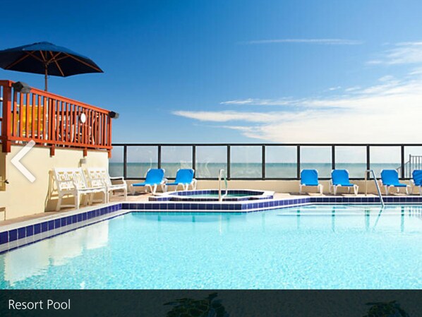 A resort pool with a clear view of the ocean, blue lounge chairs, and a red railing.