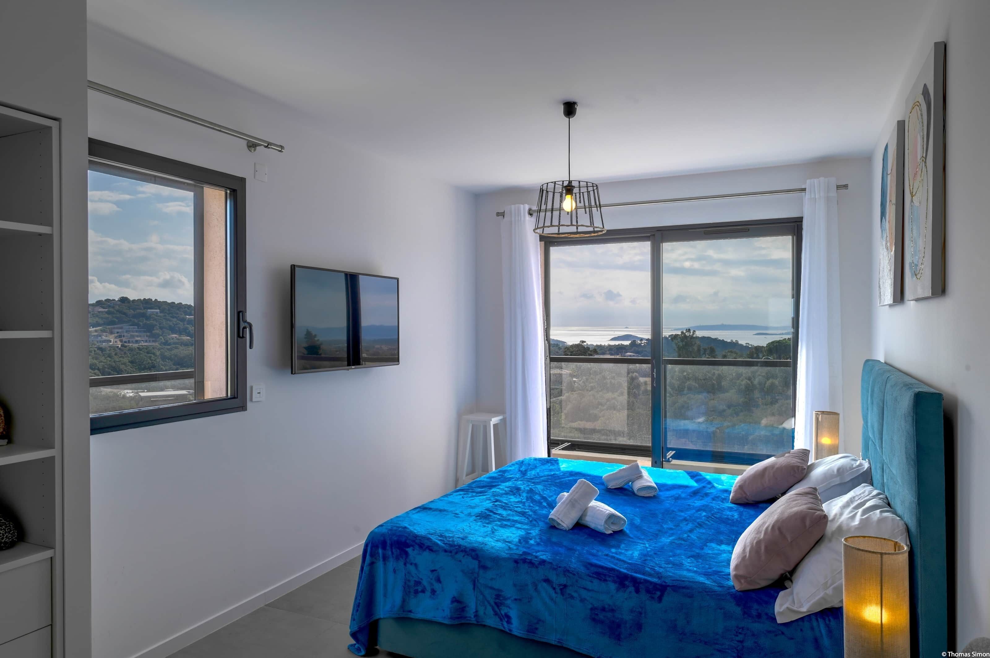 A bedroom with a large window, a bed with blue bedding, a television on the wall, and a view of the landscape.