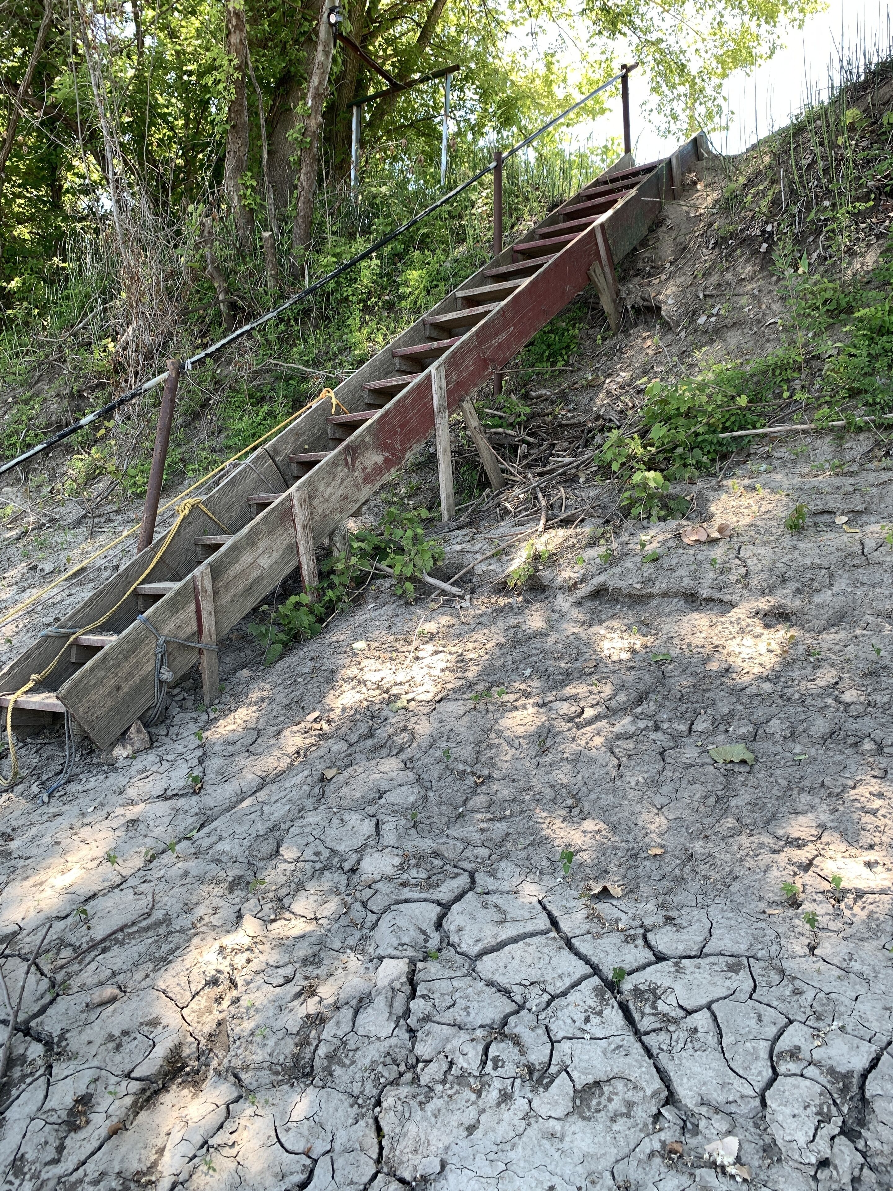 A wooden staircase with metal railings, leading up a dry, cracked ground with sparse vegetation.