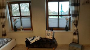 Huoneessa on kaksi ikkunaa, sänky, penkki ja pieni pöytä, jonka päällä on koristeellisia tyynyjä.