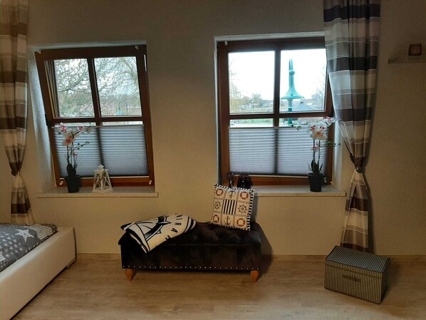 Una habitación con dos ventanas, una cama, un banco y una pequeña mesa con almohadones decorativos.