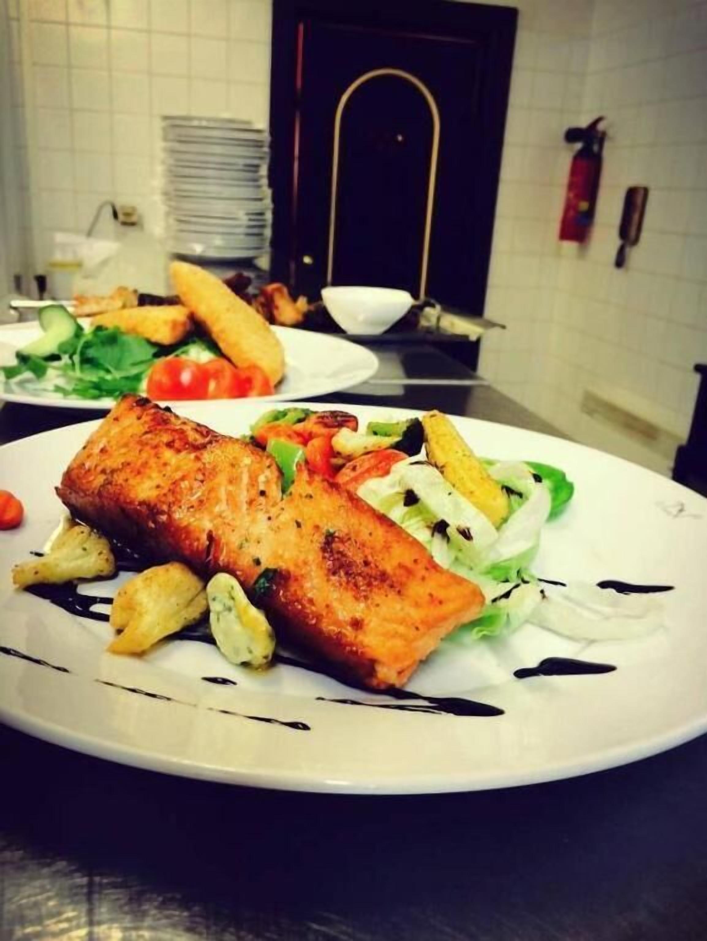A plate of grilled salmon with vegetables and a dark sauce, with a kitchen background.