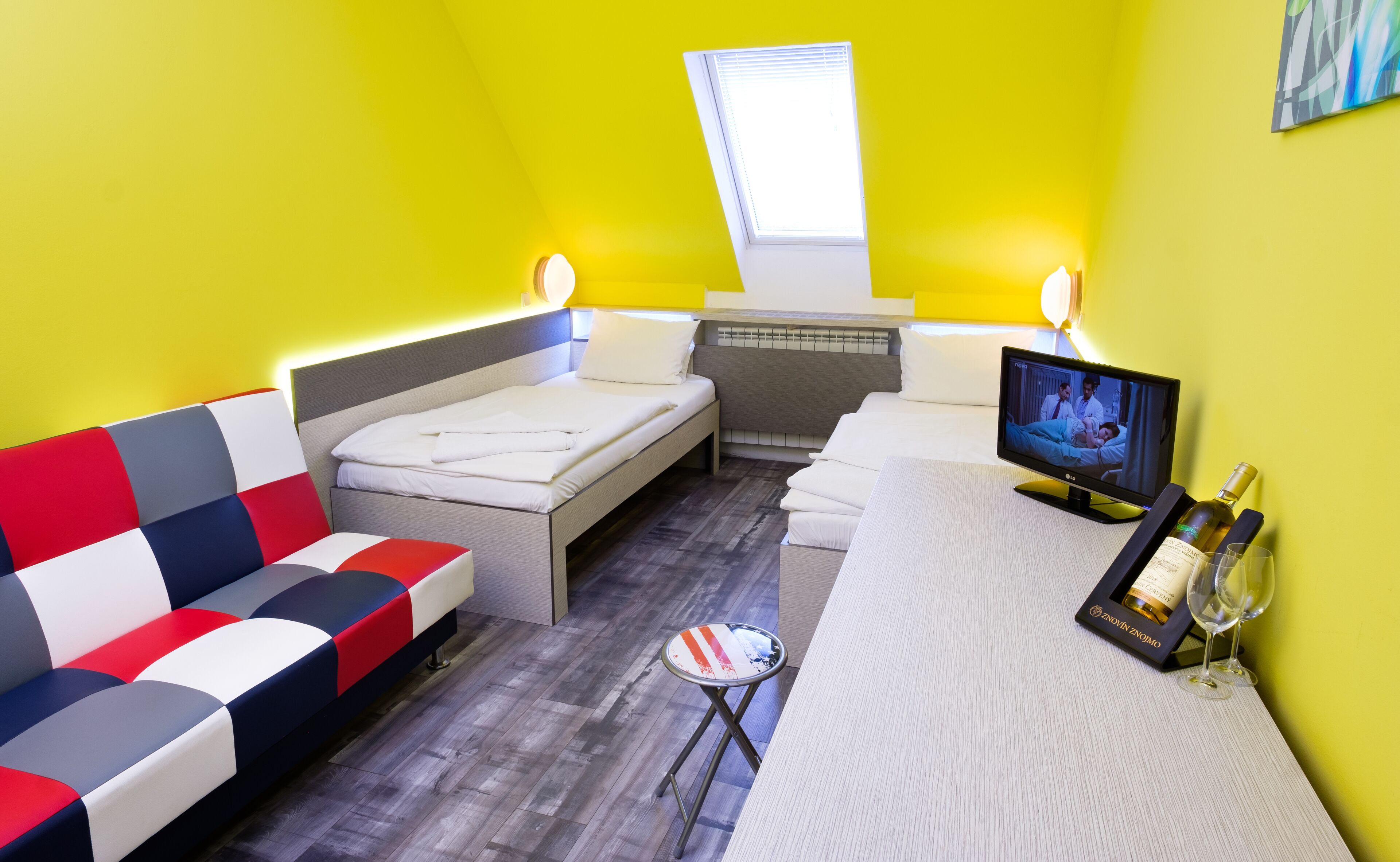 A small, brightly lit room with a bed, a sofa, a TV, and a small table with a bottle and glasses.