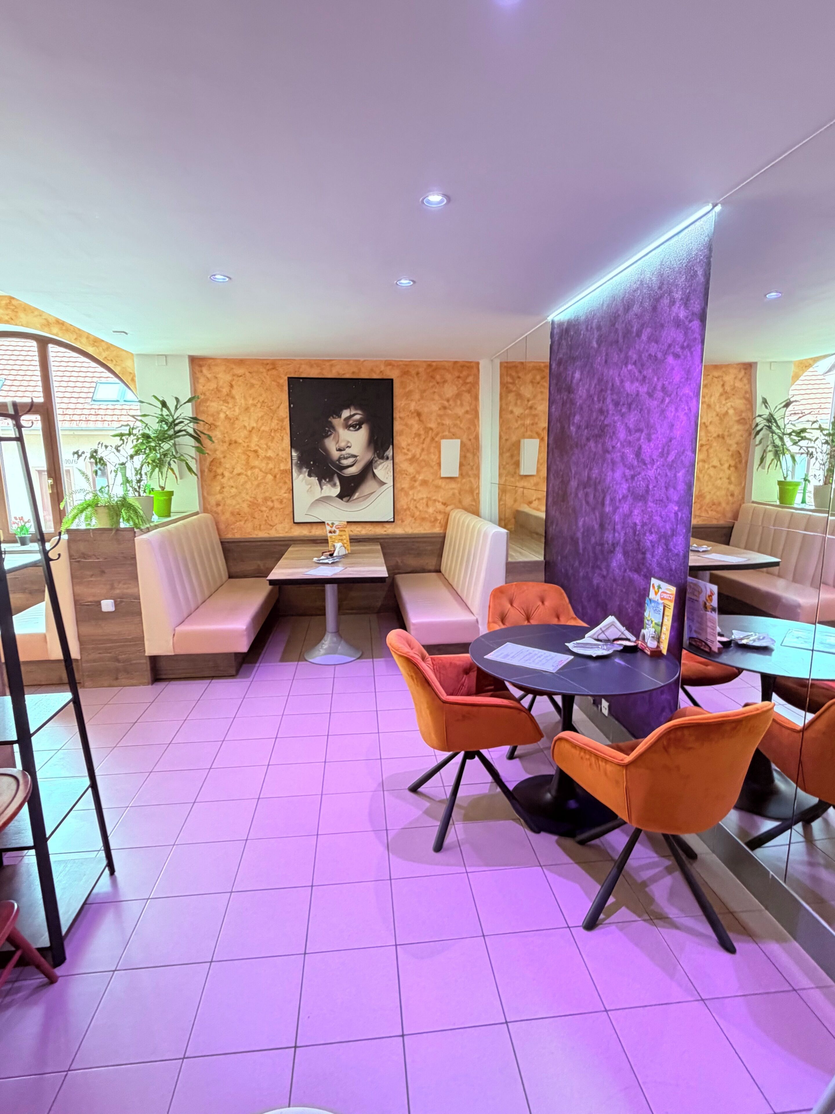 A restaurant with booths, tables, and orange chairs. There is a large painting on the wall and potted plants.