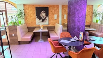 A restaurant with booths, tables, and orange chairs. There is a large painting on the wall and potted plants.