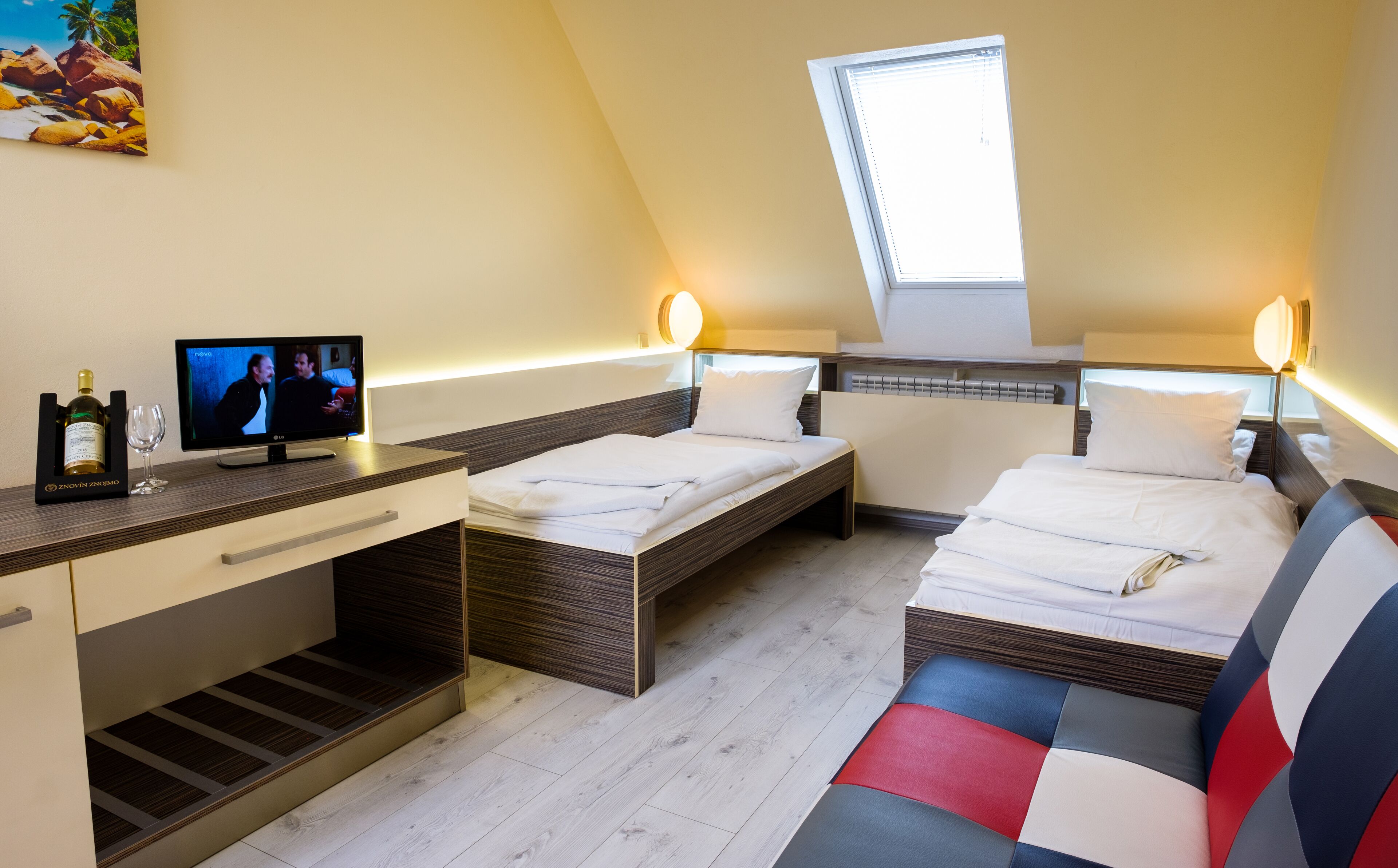 A modern hotel room with two beds, a desk with a TV, and a small window.