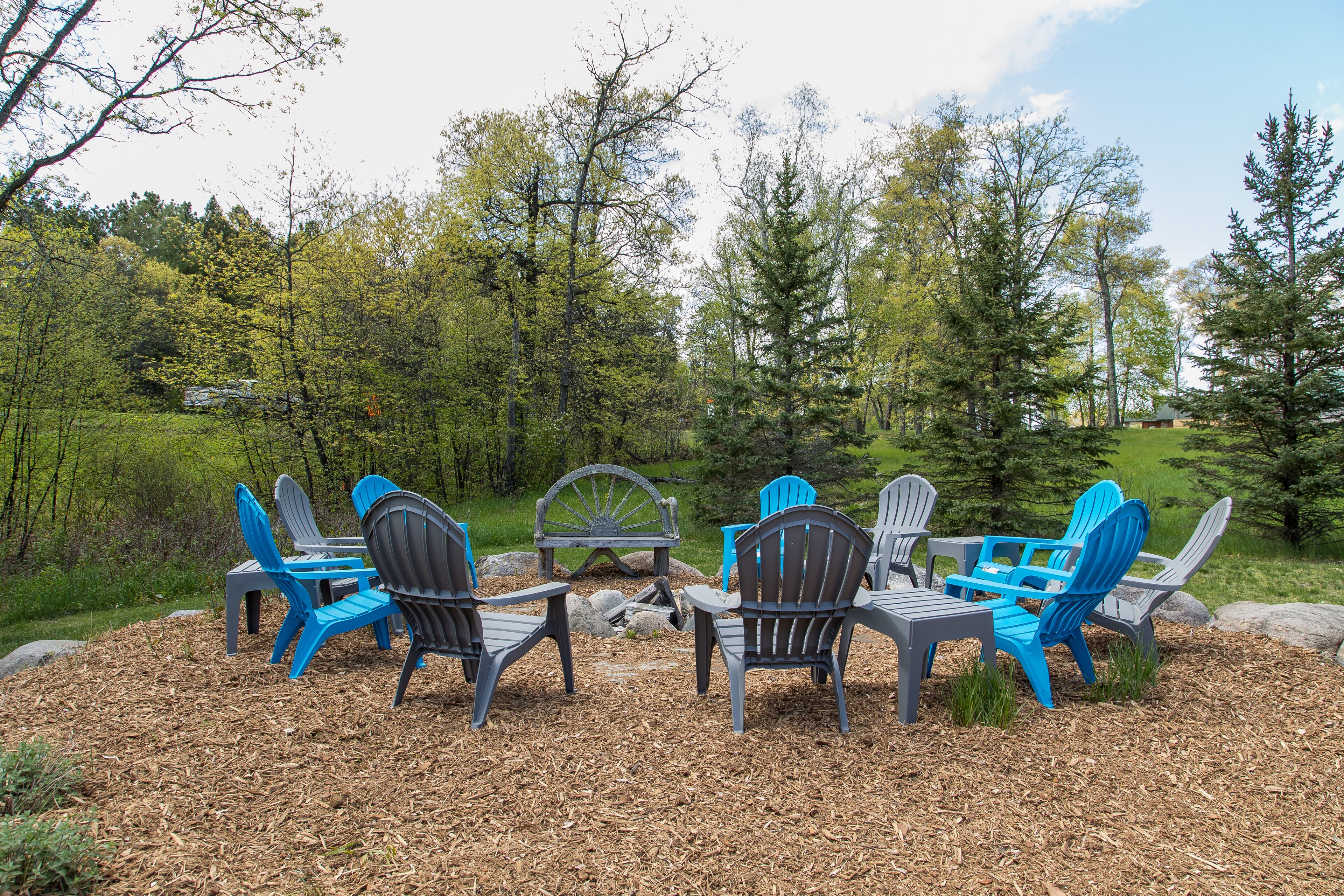 Un espace extérieur aménagé avec des chaises Adirondack et une table, entouré d’arbres et d’un champ d’herbe.