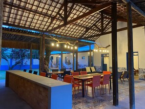 Un área de comedor al aire libre con techo de paja, luces colgantes y vista al mar.