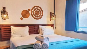 Habitación de hotel con techo de madera, una cama con ropa blanca, dos lámparas de pared y una ventana con contraventanas azules.