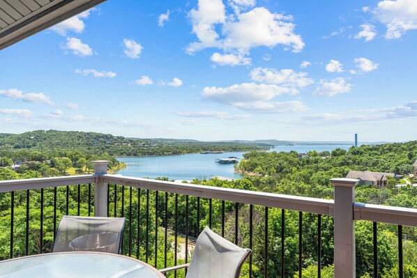 A balcony with a table and chairs overlooking a lake, surrounded by greenery.