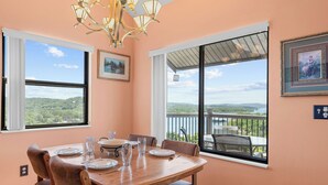 Un coin repas avec une table dressée pour quatre personnes, de grandes fenêtres donnant sur un lac et des collines, et un lustre au-dessus.