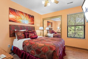 A bedroom with a large bed, a ceiling fan, a window with blinds, and artwork on the walls.