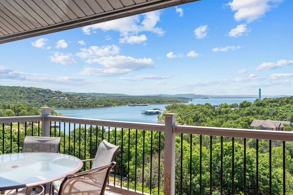 A balcony with a table and chairs, overlooking a lake with boats and a distant cityscape.