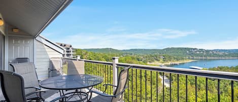 A balcony with a table and chairs, overlooking a lake and forested hills.