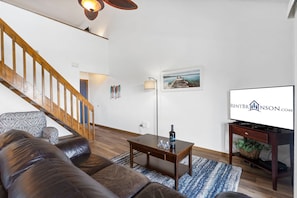A living room with a wooden staircase, a sofa, a coffee table, a floor lamp, a television, and a framed picture on the wall.