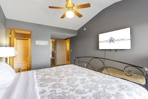 A bedroom with a bed, a ceiling fan, a TV mounted on the wall, and a door leading to another room.