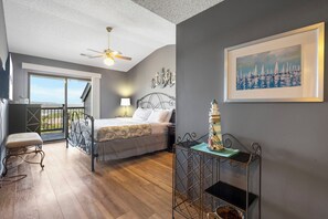 Un dormitorio con cama, escritorio, silla, balcón y un cuadro.