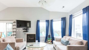 A modern living room with beige sofas, a glass coffee table, a fireplace, and a flat-screen TV mounted on the wall. - Short walk to Beach with Pool, Game Room, and Spa! (Corolla)