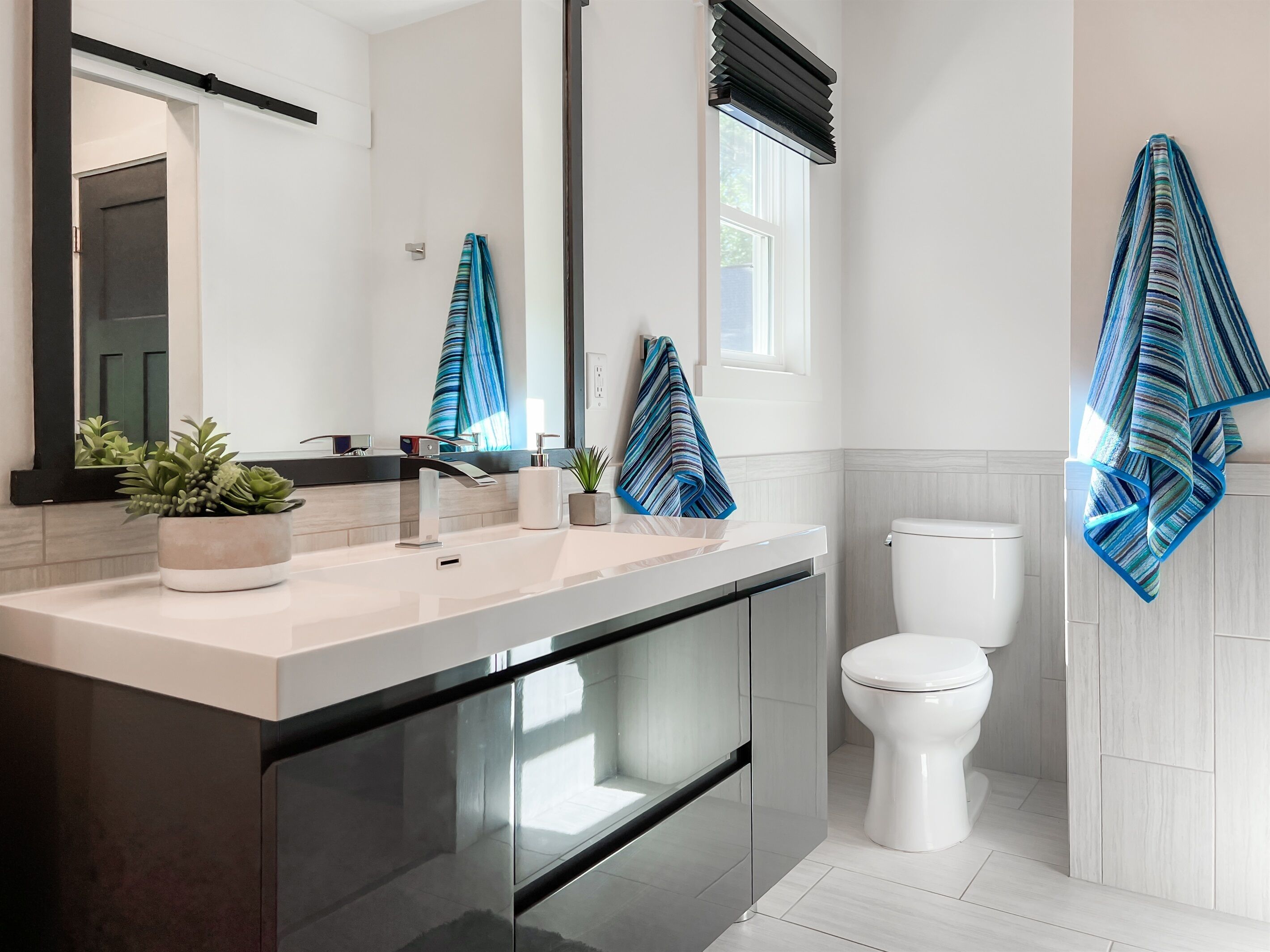 A modern bathroom with a large mirror, a sink with a faucet, a toilet, and a window with a black blind. There are two towels hanging on the wall and a potted plant on the counter.