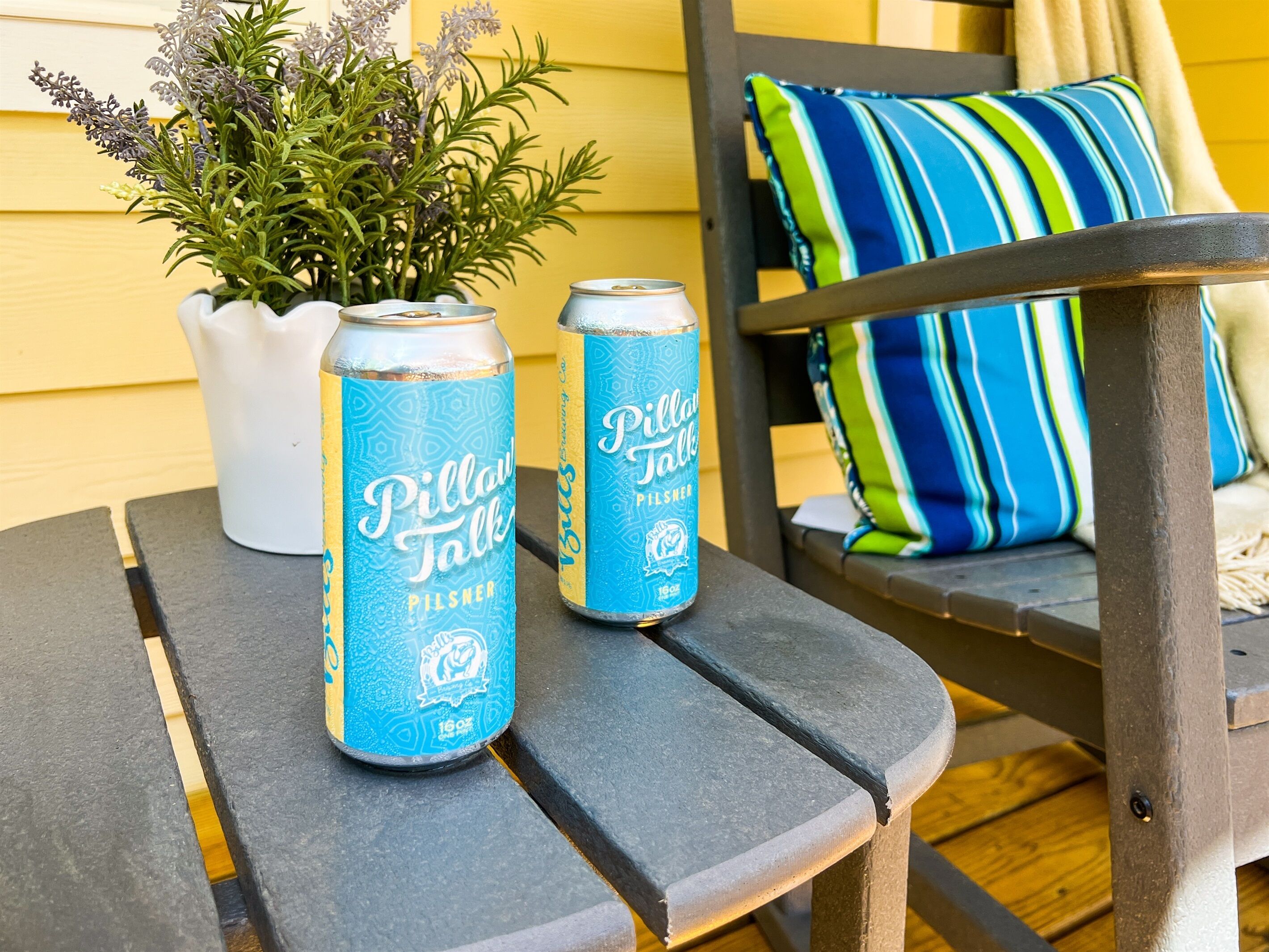 Two cans of Pillar Talk Pilsner beer on a wooden table with a potted plant and a striped pillow in the background.