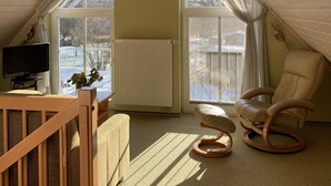 Et hyggeligt soveværelse under skrå loft med et træskrivebord, en behagelig stol, en radiator og store vinduer med udsigt over landskabet.
