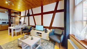 Een gezellige woonkamer met een houten plafond, een eethoek met witte stoelen, een televisie en een klein tafeltje met een klok.