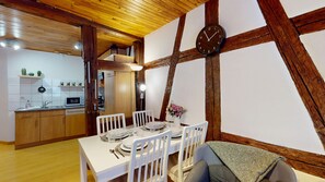 Uma sala de jantar com uma mesa branca posta para quatro pessoas, vigas de madeira no teto e um relógio na parede.