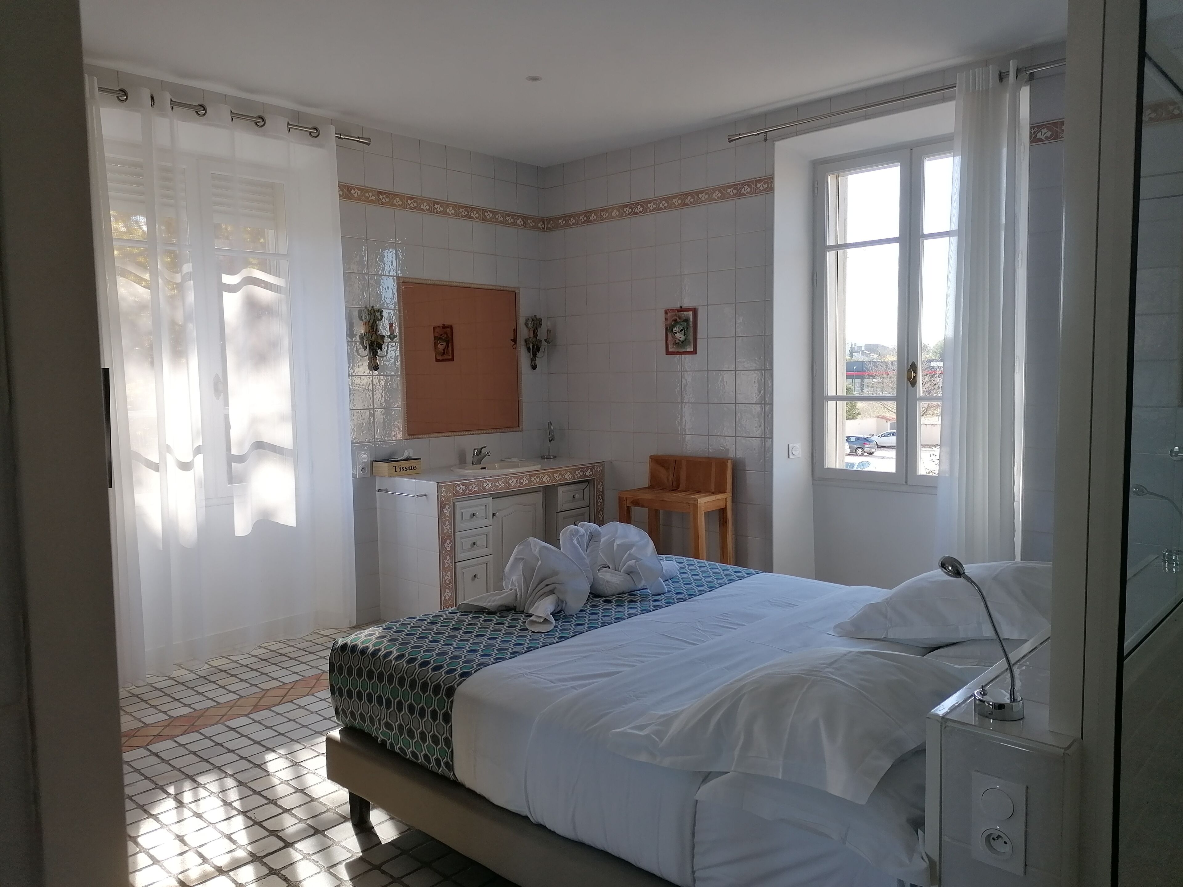 A bedroom with a bed, a bedside table, a mirror, a window, and a bathroom with a sink and a chair.