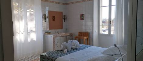 A bedroom with a bed, a bedside table, a mirror, a window, and a bathroom with a sink and a chair.