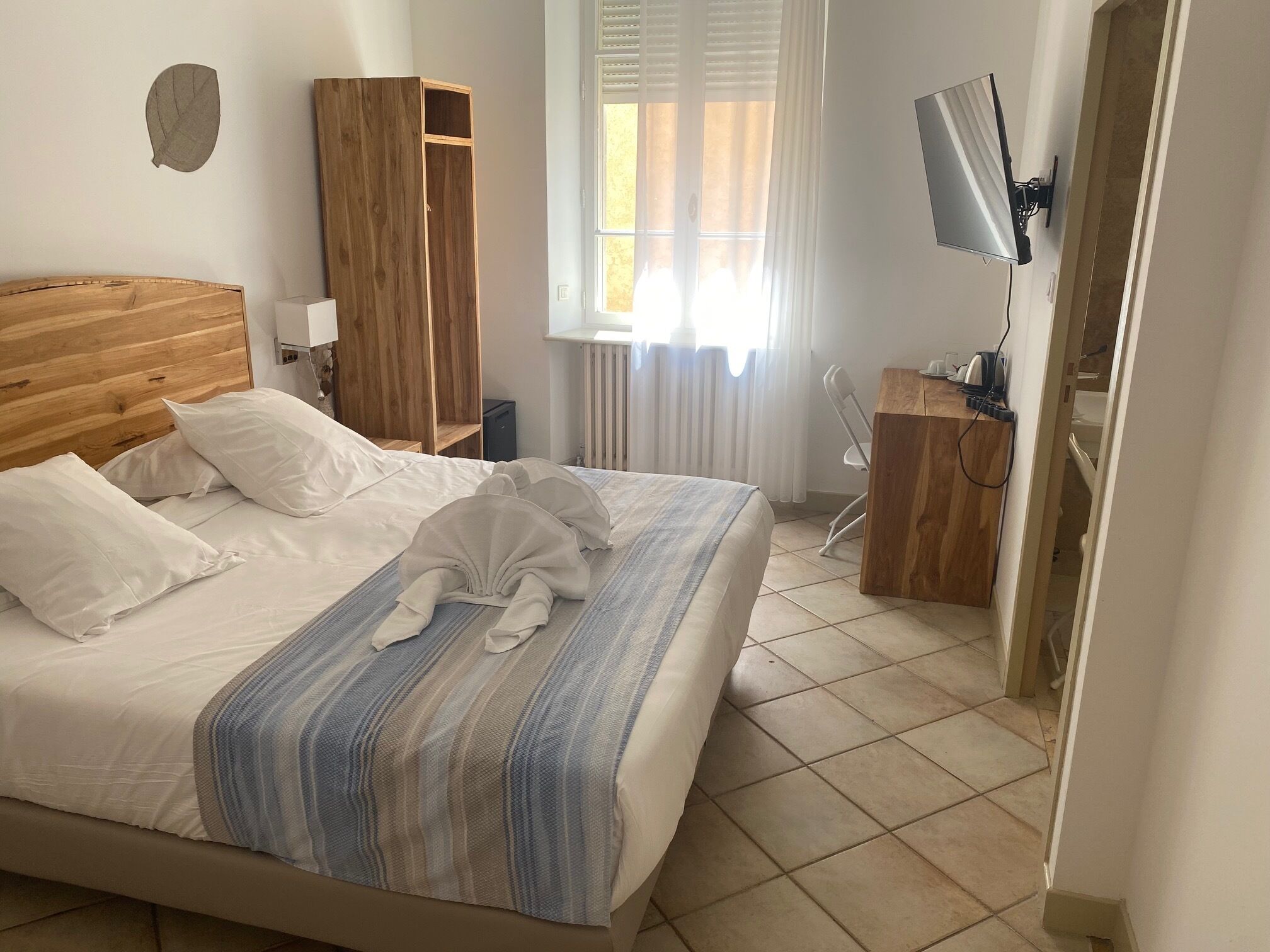 A hotel room with a wooden headboard, a bed with white linens and a striped blanket, a wooden wardrobe, a desk with a chair, a television, and a window with curtains.