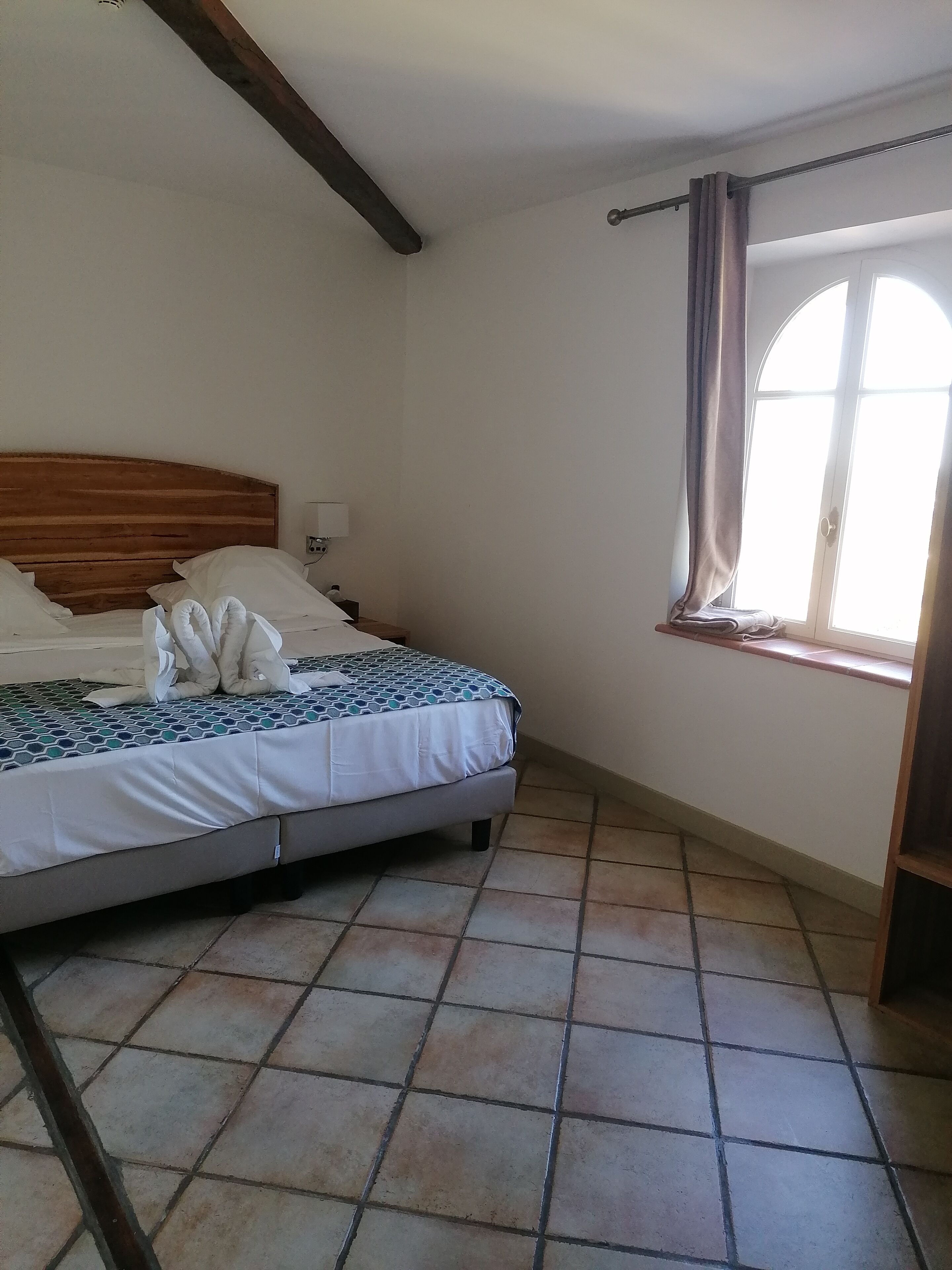 Une chambre à coucher avec un lit, une table de chevet, une fenêtre avec des rideaux et un plancher carrelé.