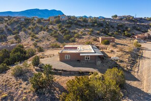 A remote, desert-style building with a flat roof and a small porch, surrounded by sparse vegetation and distant mountains.