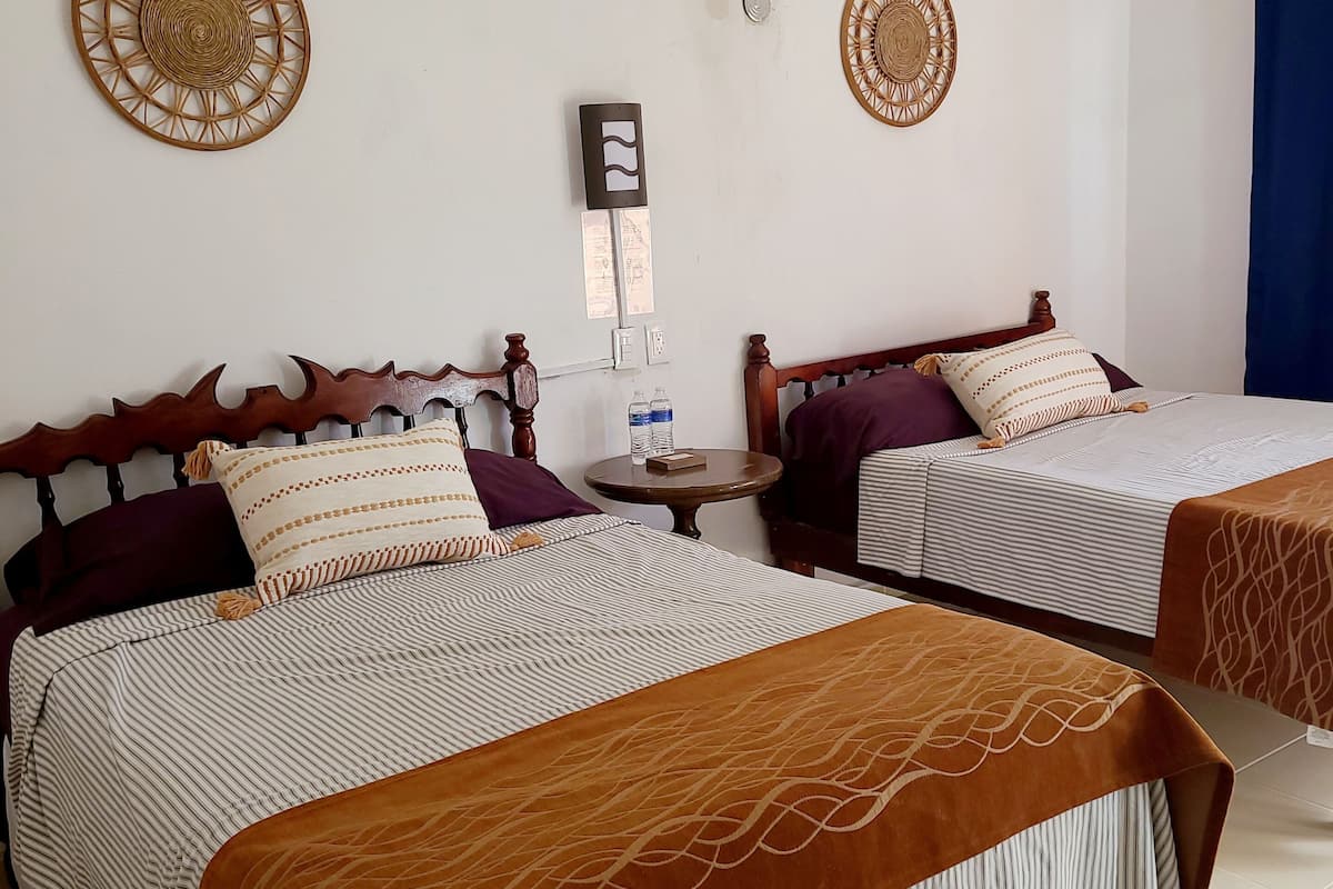 A hotel room with two beds, each with a wooden headboard, striped bedding, and a brown throw blanket. There is a small round table with a water bottle and a remote control.