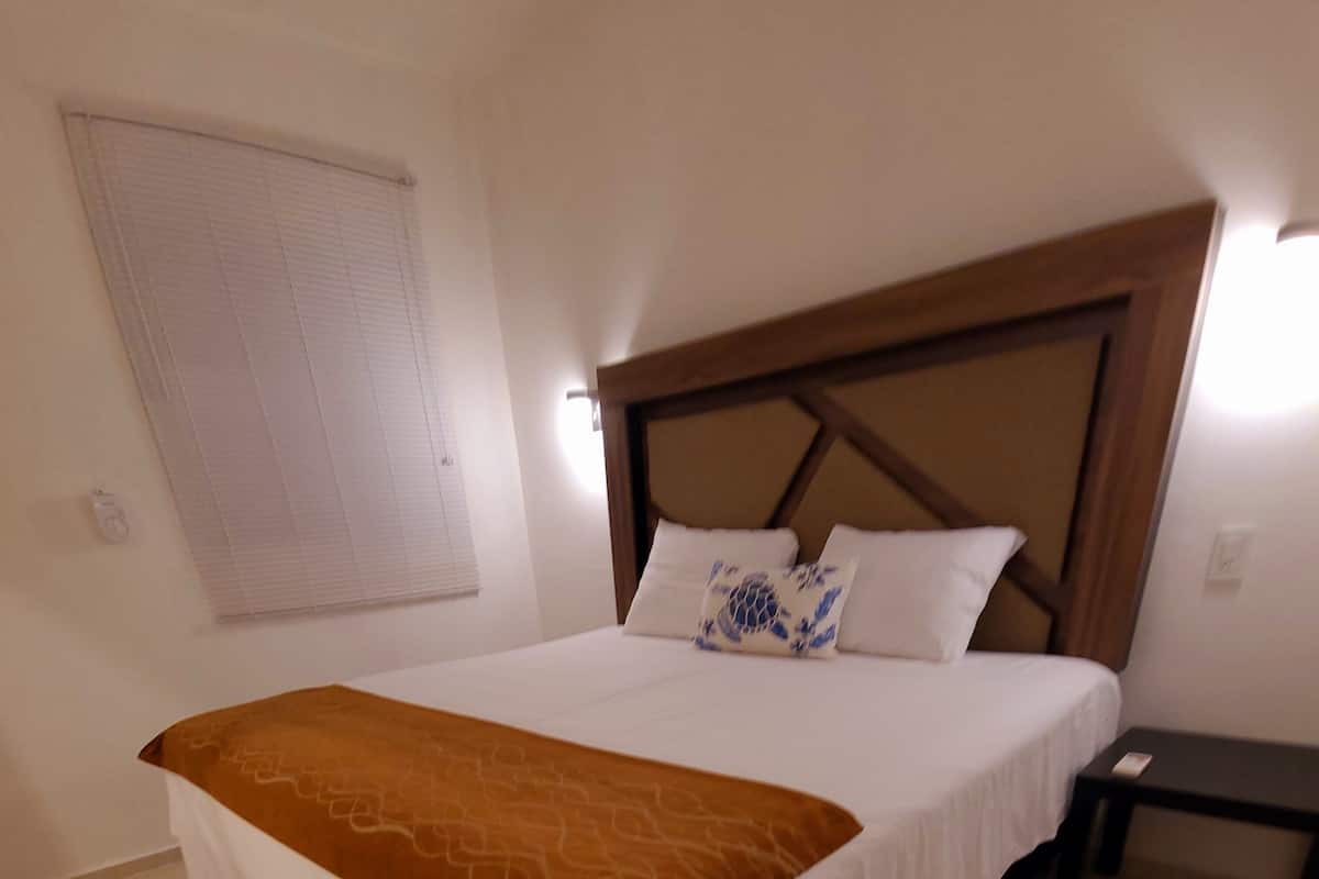 A bedroom with a large bed, a wooden headboard, a bedside table, and a window with blinds.