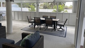 En overdækket terrasse med et bord og stole, en ventilator i loftet og udsigt til grønne områder.