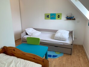 A small, neatly arranged bedroom with a single bed, a chair, a table, and a shelf with decorative items.