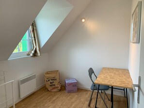 Ein kleiner, heller Raum mit schrägem Dach, einem Fenster mit Vorhängen, einem Heizkörper, einem Tisch mit Stühlen und einer großen Tasche mit dem Aufdruck 'Waschkorb'.