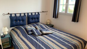 Een netjes opgemaakt bed met gestreept beddengoed, een nachtkastje met een lamp en een raam met gordijnen.