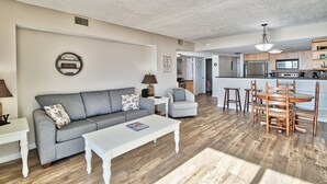 Een moderne woonkamer met een grijze bank, een witte salontafel, en een eethoek met houten stoelen. Achtergrond: een kleine keuken.