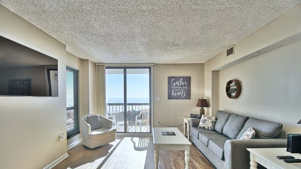 Een moderne woonkamer met een grijze bank, witte fauteuils, een klein witte tafeltje en een groot raam met uitzicht op zee.