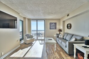 Een moderne woonkamer met een grijze bank, witte fauteuils, een klein witte tafeltje en een groot raam met uitzicht op zee.