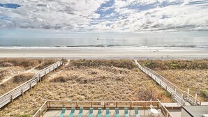 Vista de la playa con un paseo de madera que lleva a una terraza con sillas.