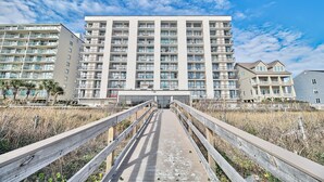 Un hotel de varios pisos con entrada central, rodeado por una pasarela de madera que conduce a una playa.