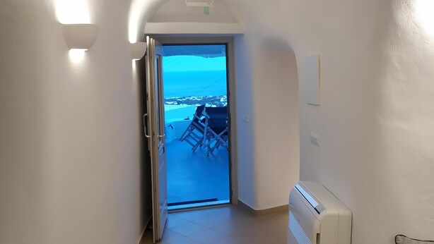 A hallway with a view of the sea through an open door, a radiator, and a chair. - Santorini Cave Suite (Thera)