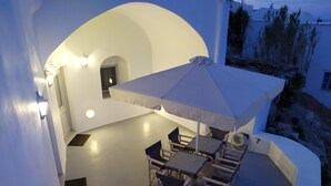 An outdoor seating area with a table and chairs under a large umbrella, surrounded by white walls and a small window. - Santorini Cave Suite (Thera)