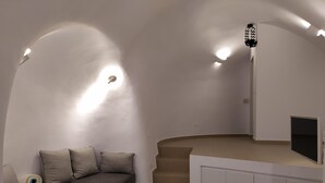A modern, minimalist living room with a curved white wall, a sofa with cushions, a small wooden coffee table, and a flat-screen TV mounted on a white cabinet. - Santorini Cave Suite (Thera)